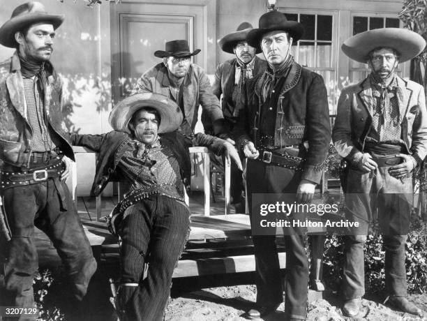 Mexican-born actor Anthony Quinn leans back against a porch railing, wearing a gunbelt and a sombrero, as actors Jack Elam , Robert Taylor and three...