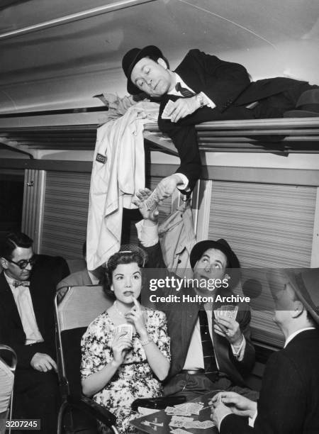American comedian Sid Caesar receives some playing cards from actor Howard Morris, lying in a luggage rack, during a card game with actors Nanette...