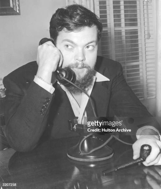 American actor and director Orson Welles seated at a table, on the telephone and holding a pipe.