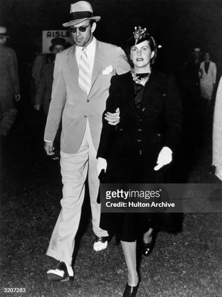 American actor Gary Cooper walking with his wife, 'Rocky', actor Sandra Shaw . He is wearing sunglasses and a hat, and she is wearing a dark suit.