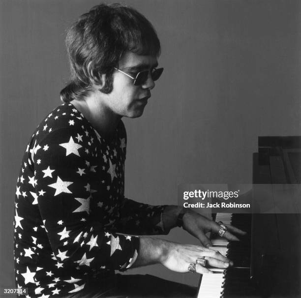 Portrait of British-born musician Elton John playing the piano and singing. John wears sunglasses, a long-sleeved shirt covered in white stars, and...