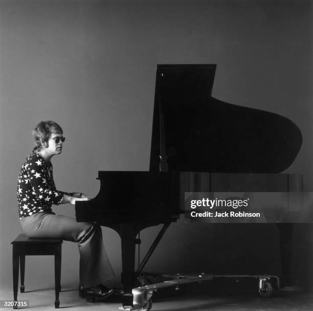 Full-length portrait of British-born musician Elton John playing a black grand piano while wearing sunglasses, a long-sleeved shirt covered in stars,...