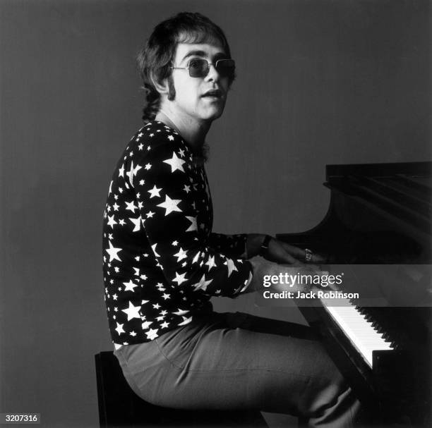 Portrait of British-born musician Elton John playing piano while wearing sunglasses and a shirt covered in stars.