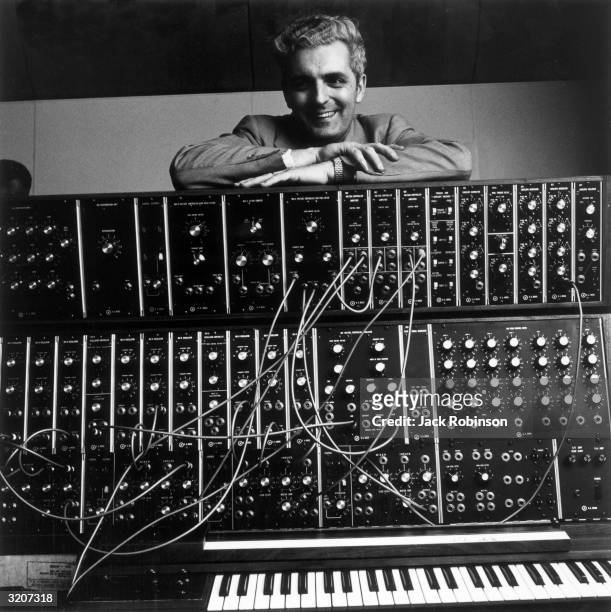 Portrait of American inventor Robert Moog smiling as he rests his arms atop his pioneering Moog synthesizer, with a keyboard and electonic circuits.