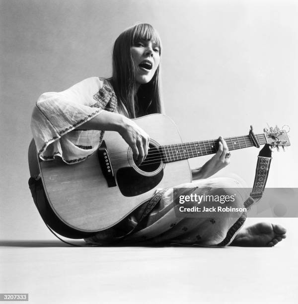 Portrait of American musician Joni Mitchell seated on the floor and playing acoustic guitar. This image was from a photo shoot for the fashion...