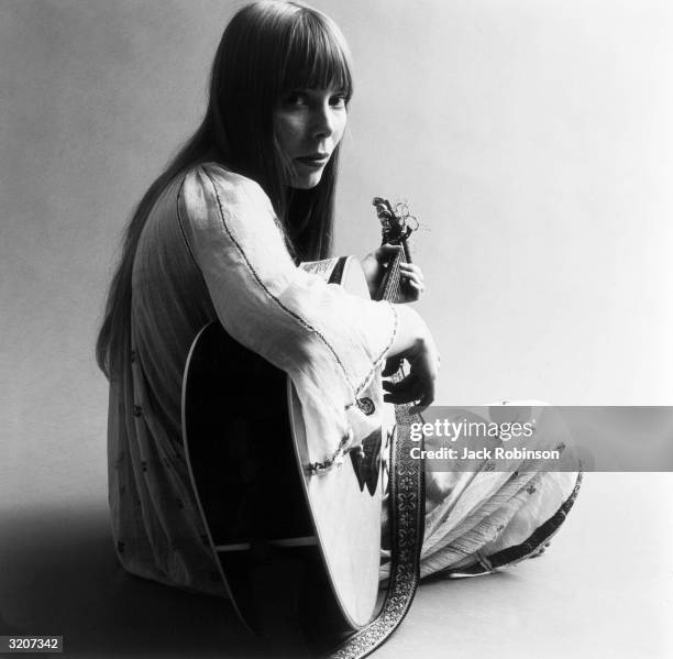 Portrait of American musician Joni Mitchell seated on the floor playing acoustic guitar. This image is from a shoot for the fashion magazine Vogue....