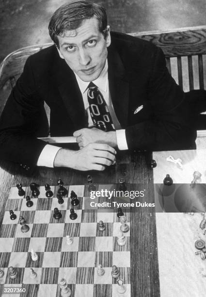 High-angle portrait of American chess champion Bobby Fischer looking up while sitting behind a chess board.