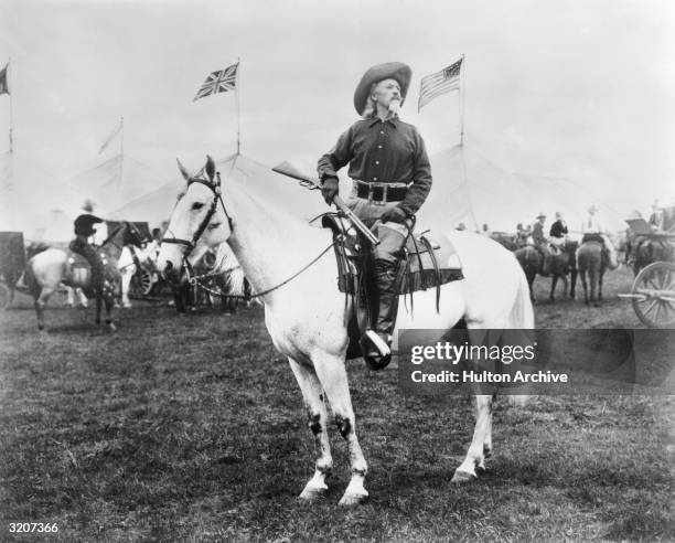 William 'Buffalo Bill' Cody American entertainer, sitting on horseback and holding a rifle, looks off into the distance as British and American flags...