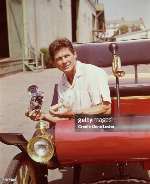 American actor Charles Bronson sits in the front of a 1920s automobile, holding a Leica flash-camera, on a studio backlot, California. He was...