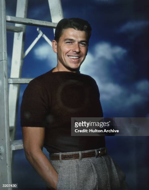 Studio portrait of American actor Ronald Reagan, wearing a short sleeve boat-neck sweater, standing near a ladder against a blue sky backdrop.