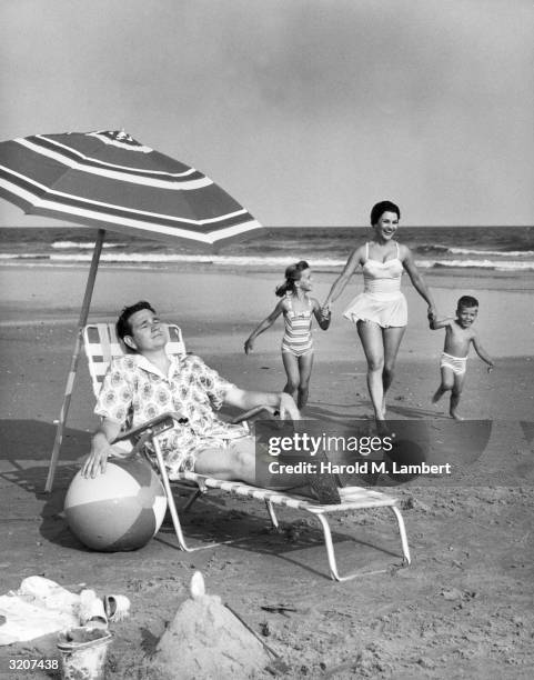 Man lounges in a deck chair under a beach umbrella, while a mother runs with their son and daughter on the sand.