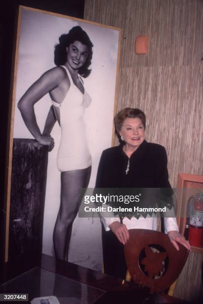 American actor and swimmer Esther Williams standing next to a full-length billboard of herself dressed in a bathing suit at a book party for her...