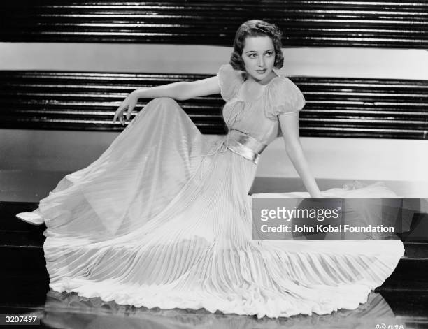 British-born actress Olivia de Havilland wearing a full-length pleated gown with a gathered waist and puffed sleeves.