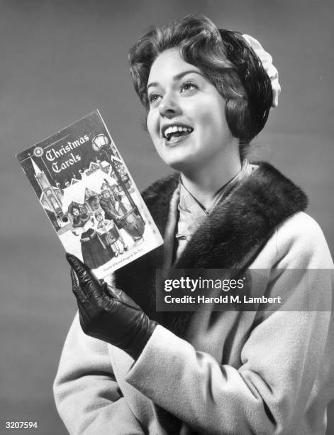 Studio portrait of a woman singing Christmas carols from a holiday song book, 1960s. She wears a winter coat and leather gloves.