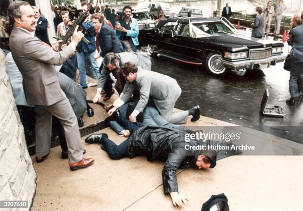 Man holding an automatic rifle, men lying on the sidewalk, members of the press, and cars at the assassination attempt on United States president...