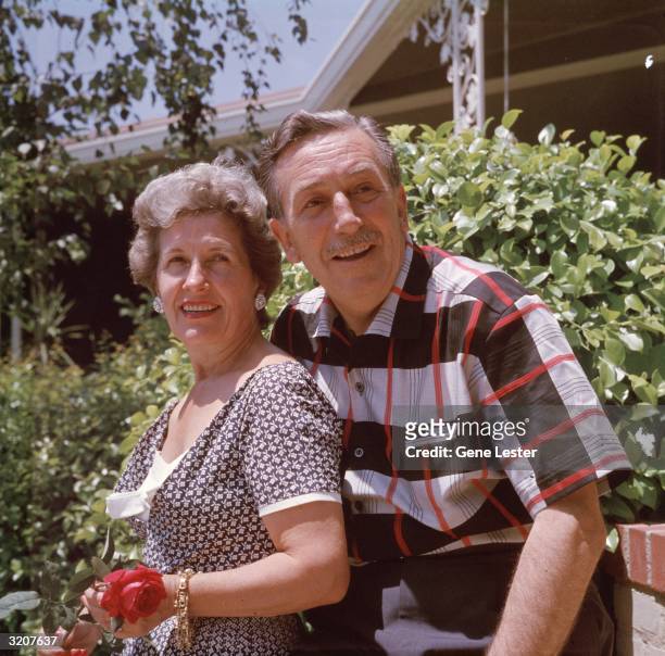 Portrait of American animator Walt Disney and his wife, Lillian, smiling while posing outdoors in front of shrubs at their home. Lillian holds a red...