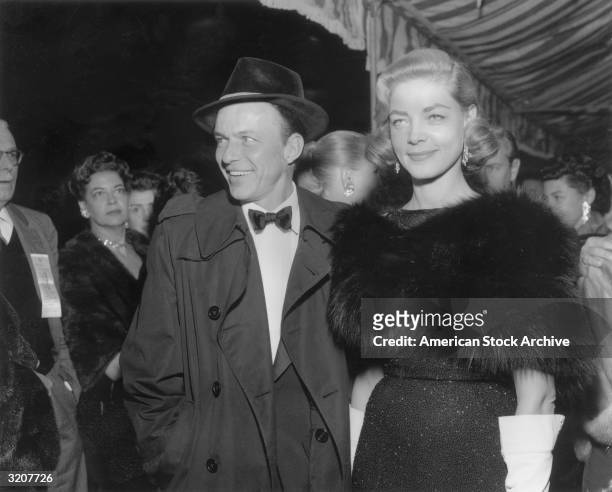 American actor and singer Frank Sinatra with American actor Lauren Bacall, smiling at a formal event. Sinatra is wearing a dark raincoat over a...