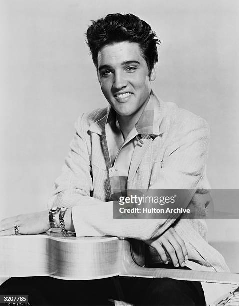 Portrait of American rock musician Elvis Presley seated with his arms crossed on top of a guitar turned downwards on his lap.