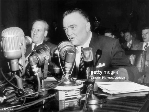 Federal Bureau of Investigations Director J Edgar Hoover sits at a table, speaking into several microphones.