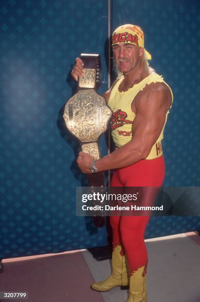 Full-length image of American wrestler Hulk Hogan displaying his championship belt. Hogan wears a costume consisting of a yellow 'HOGAN' bandana, a...