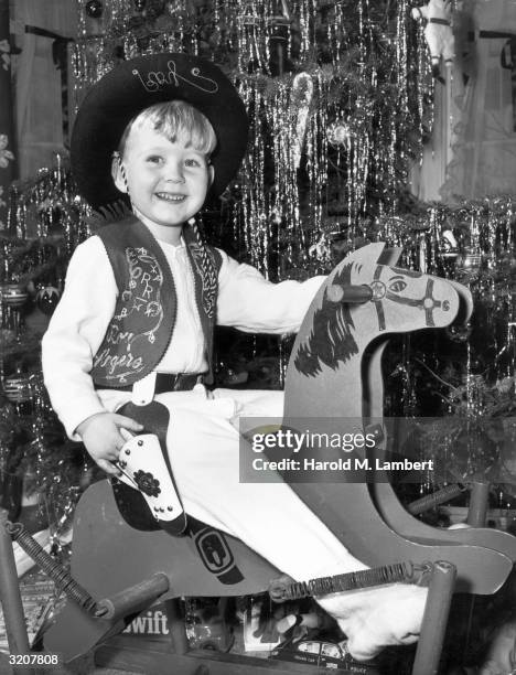 Young boy wearing a cowboy outfit smiles as he rides his new wooden rocking horse in front of the Christmas tree, 1950s. He wears a matching Roy...