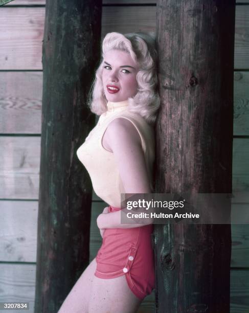 Portrait of American actor Jayne Mansfield, wearing a sleeveless sweater, pointy bra and red shorts, leaning against a wooden post, 1950s.