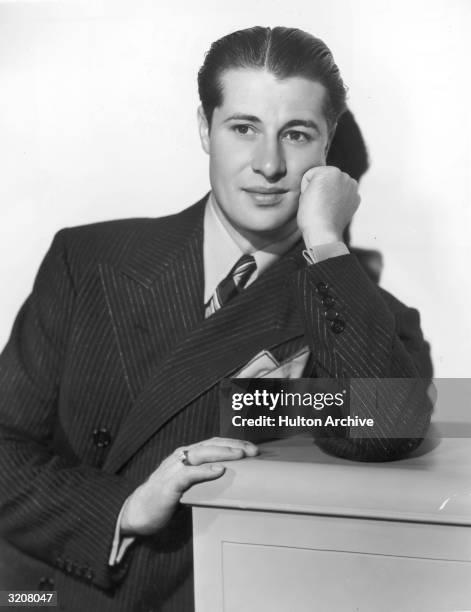 Portrait of American actor Don Ameche, wearing a double-breasted pinstriped suit with a pocket square, leaning on a cabinet.