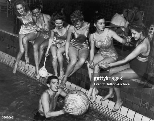 American actor Ronald Reagan grasps a beach ball in a swimming pool as six female actors wearing swimsuits sit on the edge of the pool, on board the...