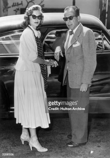 Full-length image of married American actors Lauren Bacall and Humphrey Bogart standing by an automobile. They wear sunglasses.
