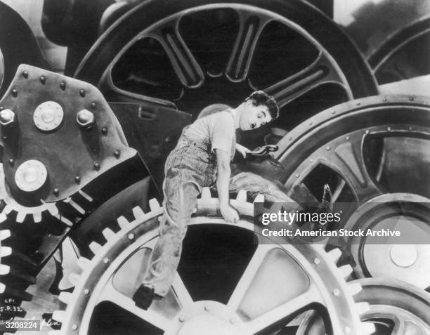 British actor and director Charles Chaplin , wearing overalls and holding a wrench, sits on an enormous set of gears in a still from Chaplin's film,...