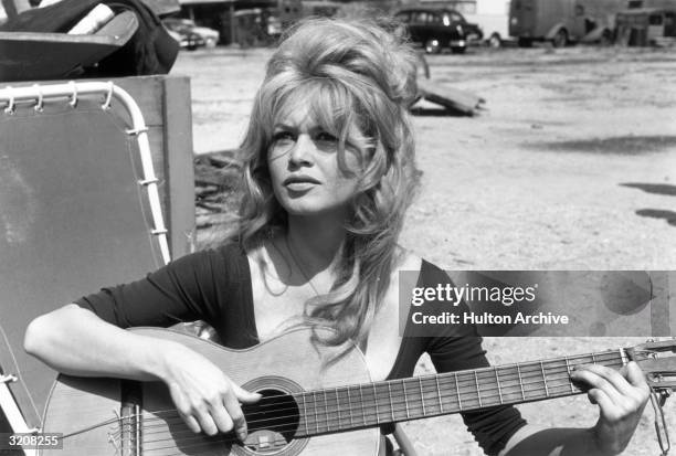 French actor Brigitte Bardot playing guitar while sitting on a lawn chair in an empty lot.