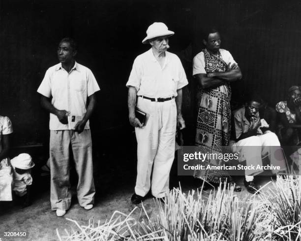 Full-length image of German missionary and physician Dr. Albert Schweitzer , wearing a pith helmet, standing outdoors with a group of people, Africa.