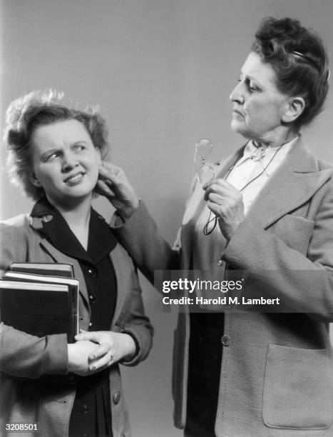 Female teacher holding a pair of spectacles pinching the ear of a female student carrying books. The student is making a face at the teacher.