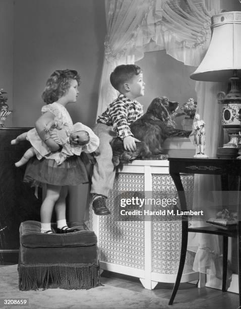 Young boy and girl with a doll look out a window with their dog.