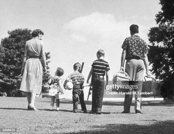 Full-length view of a family of five, including two boys and a girl, walking in a park with picnic baskets, a thermos, and toys for outdoor fun.