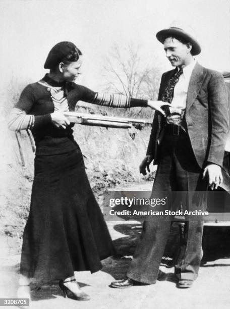American criminal Bonnie Parker aims a shotgun at her partner, Clyde Barrow while clowning beside an automobile, early 1930s.