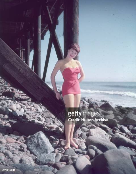 Full-length portrait of American actor Shirley MacLaine, wearing a pink strapless swimsuit, standing on the rocks beneath a wooden pier by the...