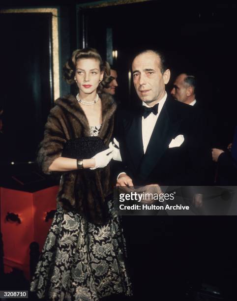 Married American actors Lauren Bacall and Humphrey Bogart stand in a foyer after arriving together. Bacall wears a brocade dress, a mink wrap, and a...