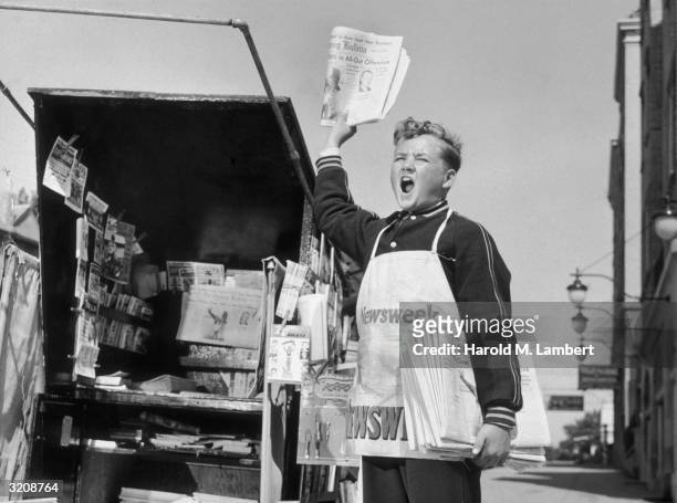 Full-length image of a newsboy shouting beside a newsstand while holding a newspaper in the air and a bundle of 'The Evening Bulletin' newspapers...