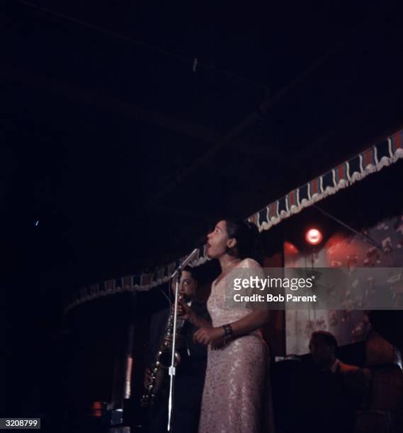 American jazz singer Billie Holiday performs on stage, backed by a saxophonist and a drummer, at the Sugar Hill nightclub, Newark, New Jersey.