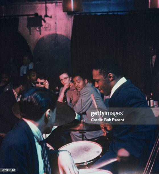 Side view of American jazz drummer and bandleader Art Blakey playing the drums as audience members watch from a table during a nightclub performance.