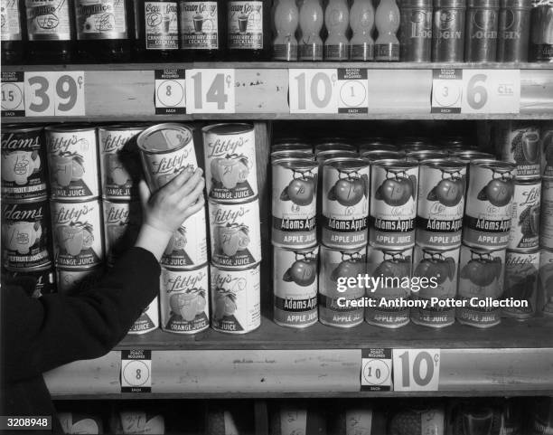 Someone reaches for a can of orange juice from a grocery shelf during World War II. Cards indicating 'Ration Points Required' for various canned...