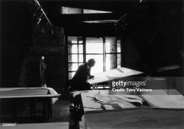 American architect Frank Lloyd Wright working on designs while sitting in front of a drafting table in his studio at Taliesin West, Arizona.