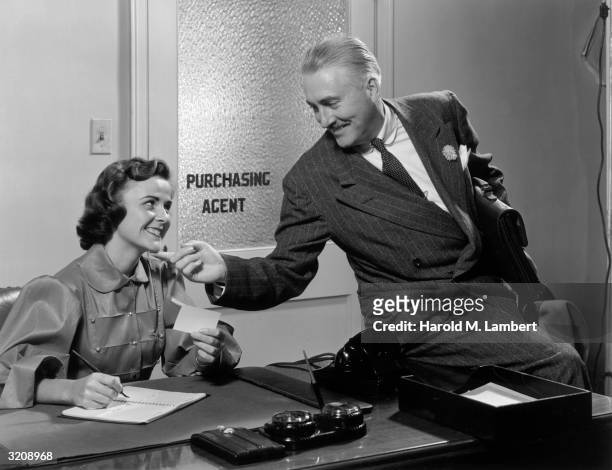 Businessman holding a briefcase smiles and places his finger under the chin of a female secretary working at her desk.