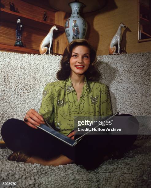 American actor Lauren Bacall smiling and sitting cross-legged on a sofa, an open book in her lap.