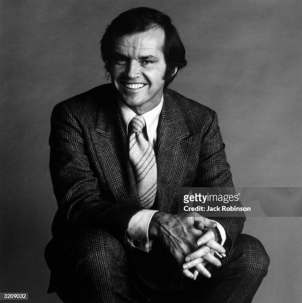 Studio portrait of American actor Jack Nicholson, crouching and smiling with his fingers intertwined.