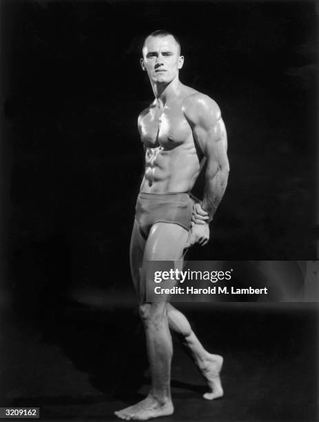 Full-length studio portrait of a male body builder posing sideways with both arms behind his back.