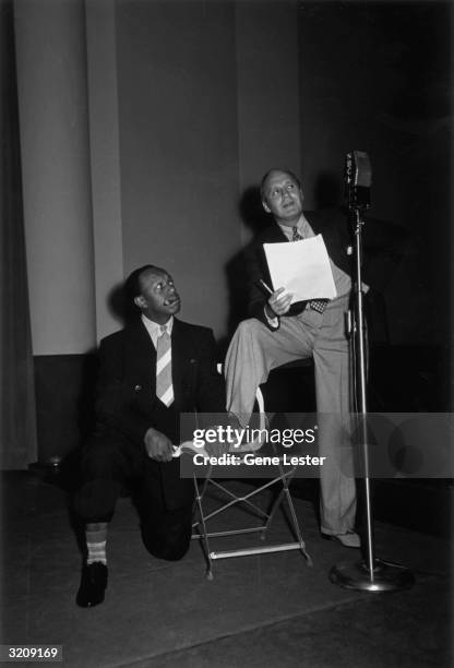 American comedian Jack Benny stands at an NBC microphone with a script as Rochester shines his shoe and smokes a cigar.