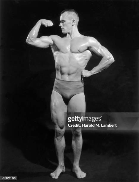 Full-length studio portrait of a male body builder flexing his right arm and looking at his biceps.