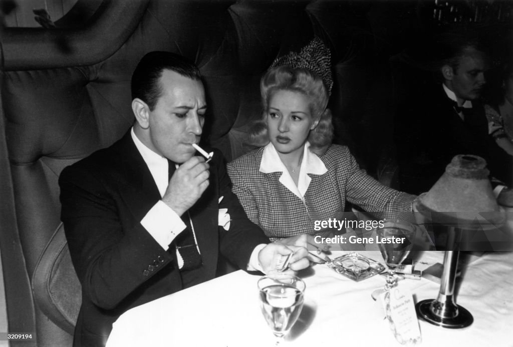 American actors Betty Grable and George Raft smoke cigarettes at a ...